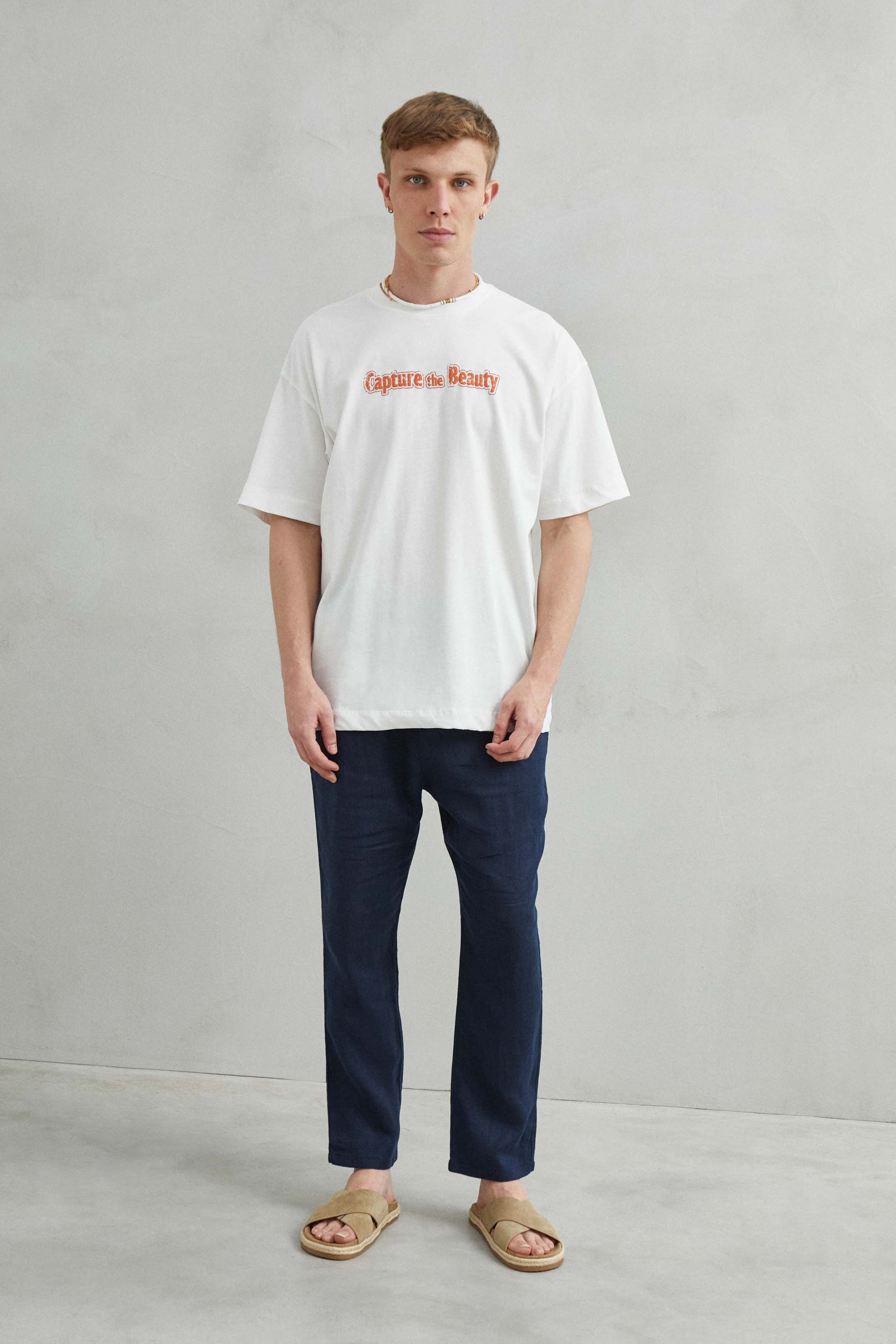 Modelo usando camiseta blanca oversize unisex con diseño montañoso, árboles, casas y mar en la espalda.