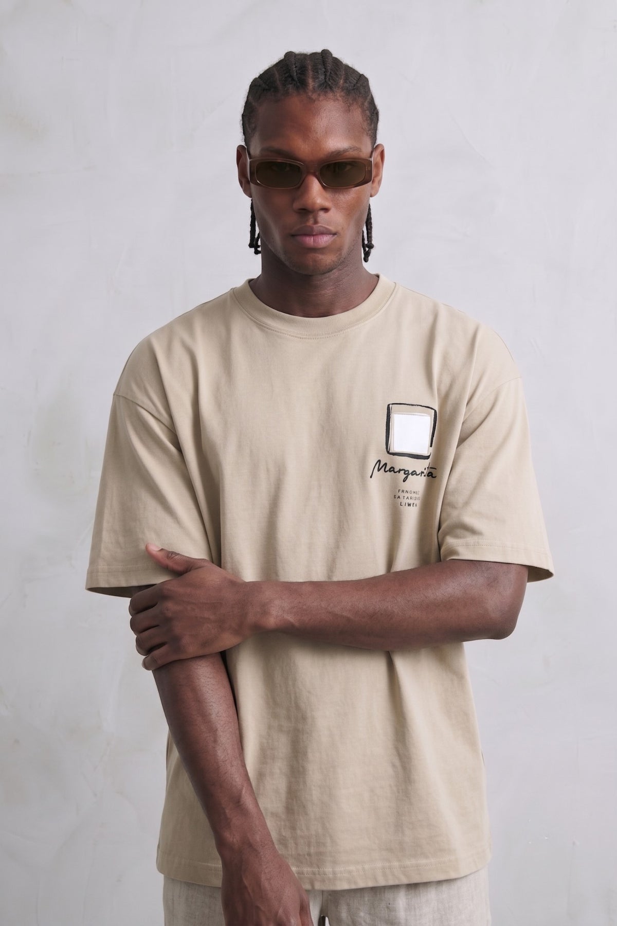 Camiseta oversize beige con pequeño cuadrado blanco de marco negro y texto manuscrito en el pecho, vista frontal.