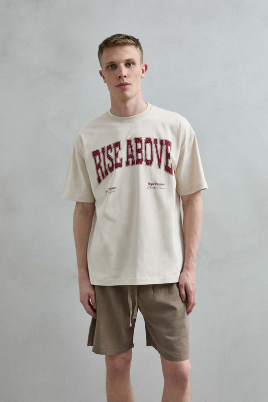 Vista frontal de medio cuerpo de la camiseta oversize beige Rise Above para hombre, con letras grandes en color burdeos.
