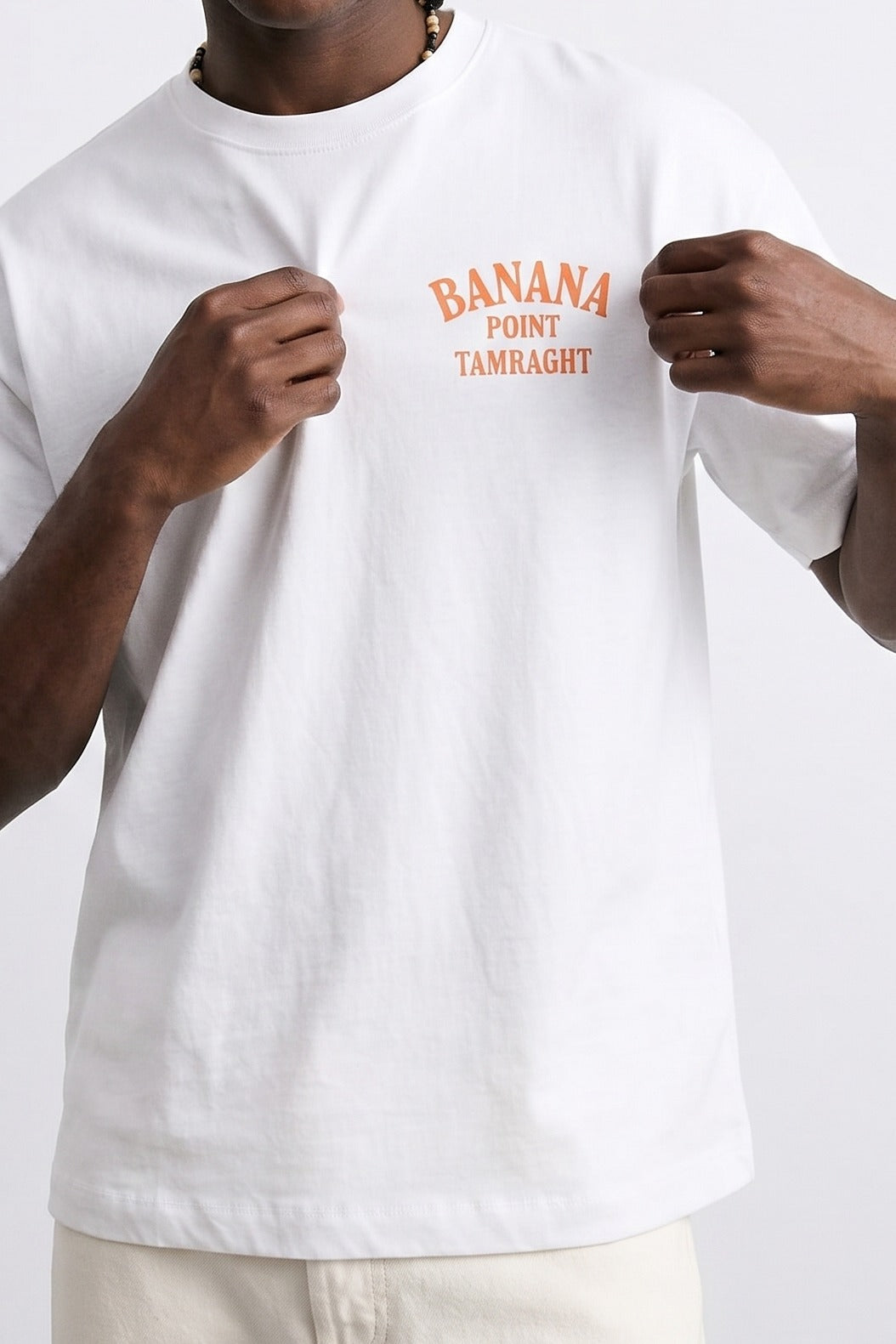 Detalle del texto naranja Banana Point Tamraght en el pecho de camiseta blanca.