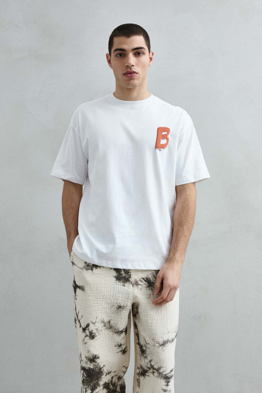 Camiseta oversize blanca BRUNO Surfer Club con letra B naranja en el pecho, vista frontal de medio cuerpo.