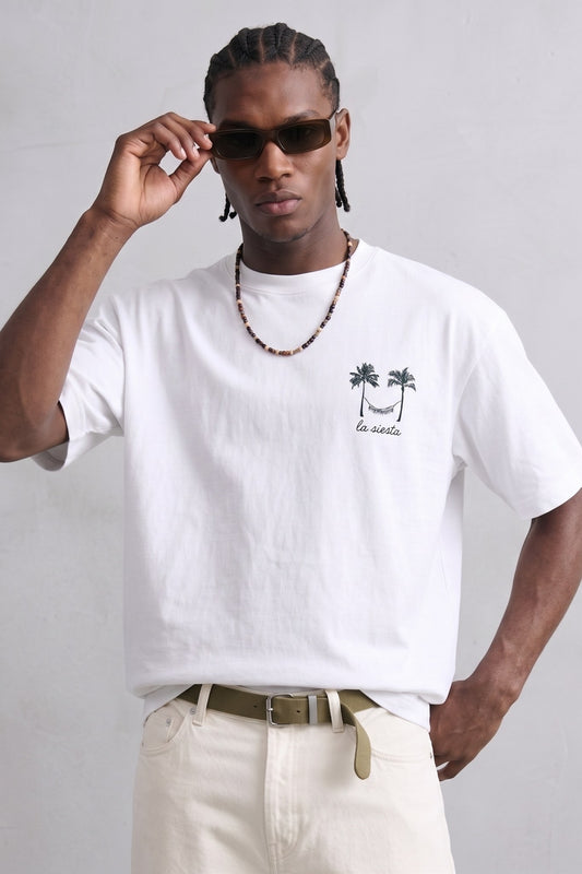 Camiseta oversize blanca La Siesta con pequeño dibujo negro de palmeras y hamaca en el pecho, vista frontal.