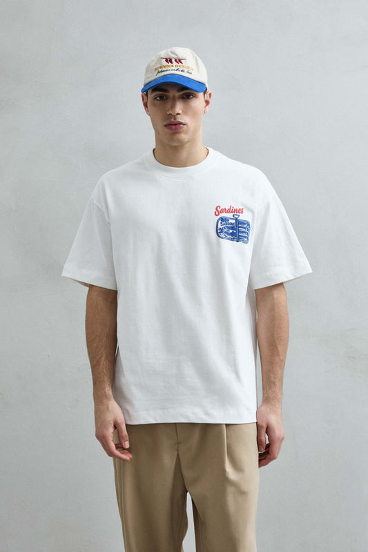 Vista frontal de medio cuerpo de la camiseta oversize blanca Sardines para hombre, con pequeño dibujo azul de una lata de sardinas y letras rojas en el pecho.