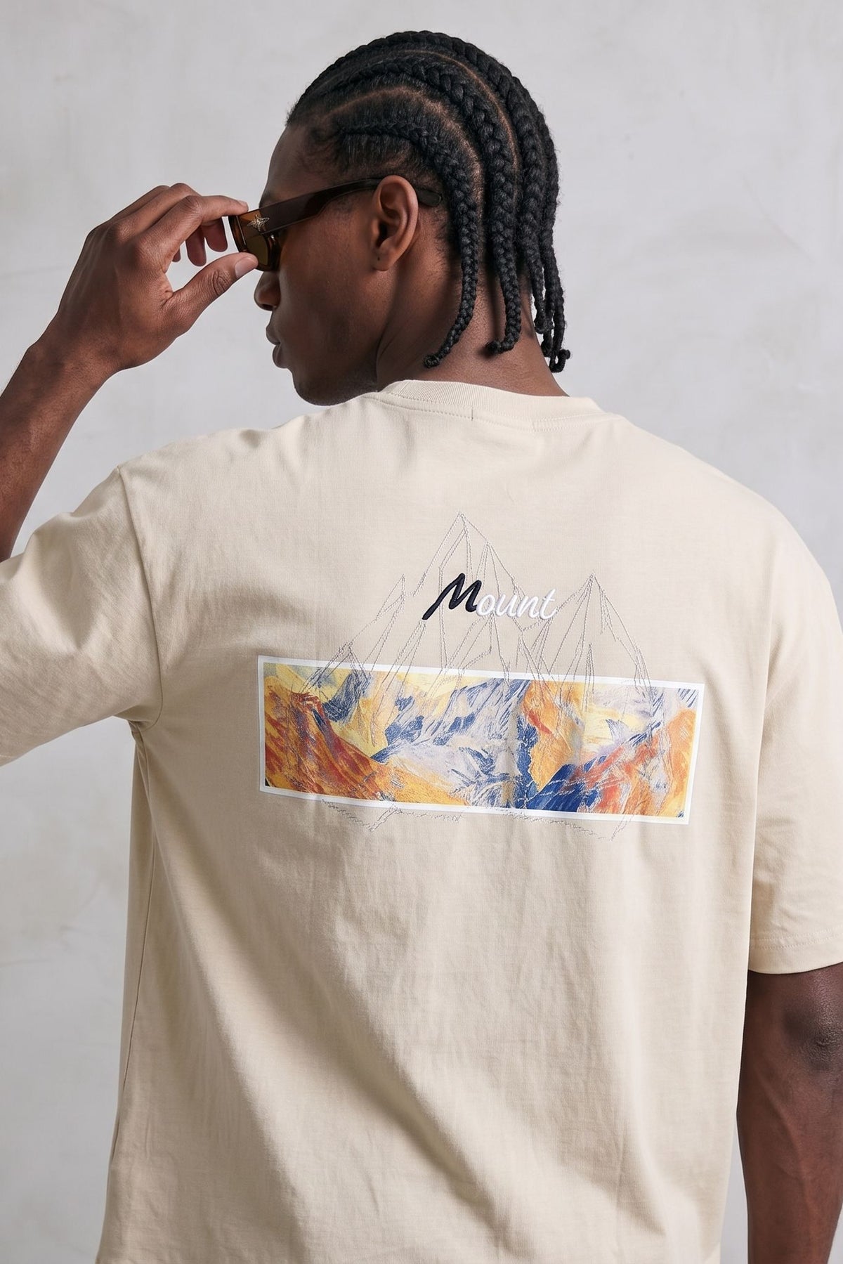 Espalda de camiseta crema con ilustración rectangular multicolor y líneas de montañas.