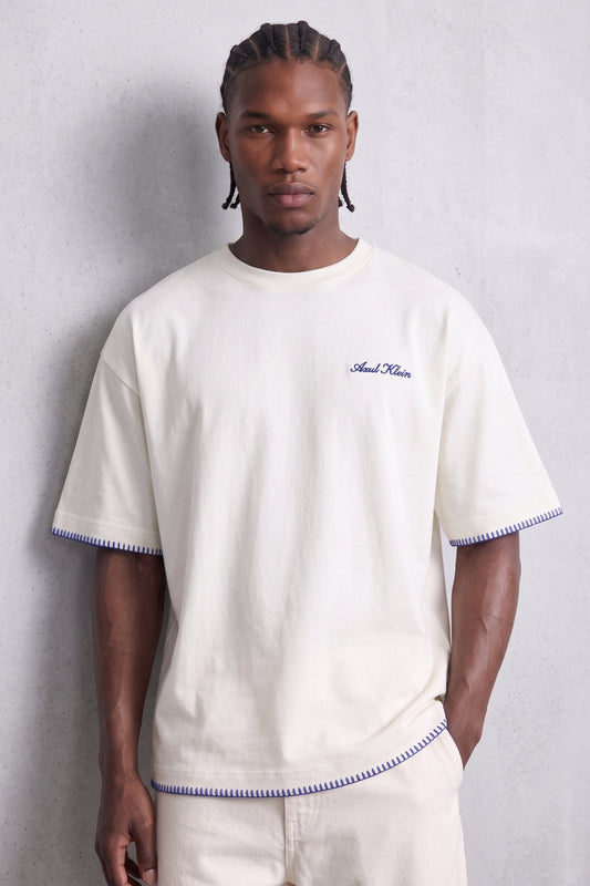 Camiseta oversize color crema con pequeño texto en el pecho y pespuntes azules en mangas y bajo, vista frontal.