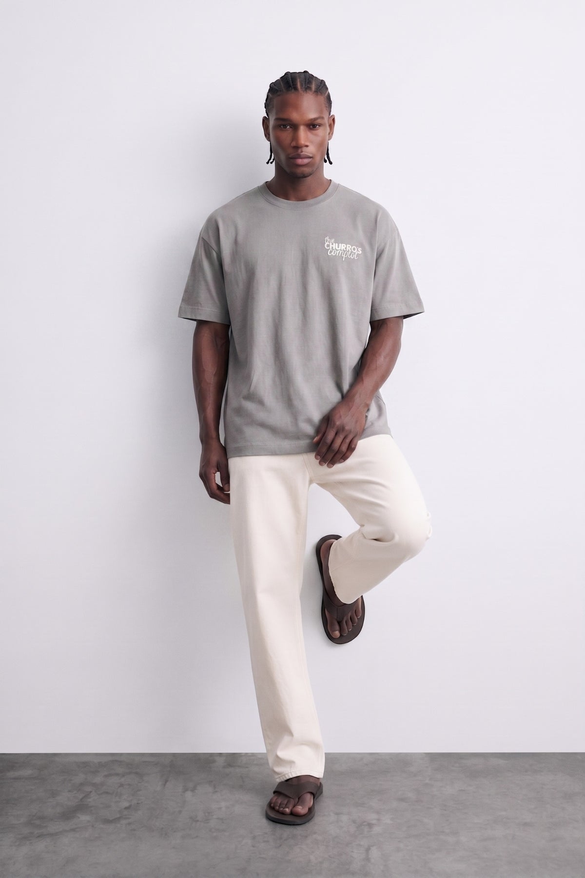 Camiseta oversize gris The Churros Complot, vista de conjunto con el modelo apoyado en una pared.