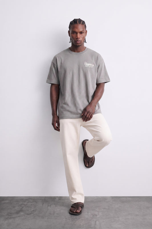 Camiseta oversize gris The Churros Complot, vista de conjunto con el modelo apoyado en una pared.