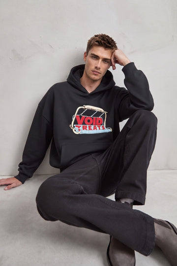 Sudadera negra oversize con capucha y estampado frontal, modelo sentado